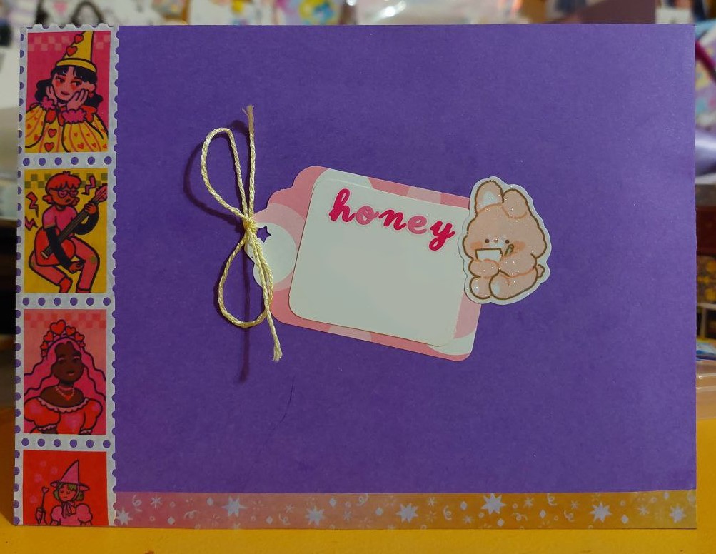 a photo of the front of a purple envelope. there is a pink gift tag in the center with the name 'honey' on it in dark pink and light pink alternating letters. a bear with a notepad overlaps the tag slightly on the right side. the left of the envelope has washi tape with various women in cute outfits.