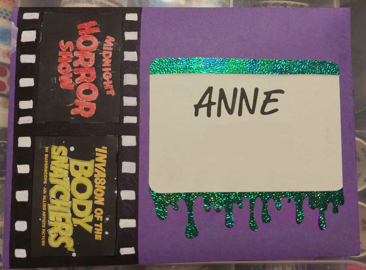 photo of the front of an envelope. there is a handmade film strip running down the left side of the purple envelope with old horror movie title cards. there is a label on the front that reads Anne and has iridescent green slime dripping from the bottom of it