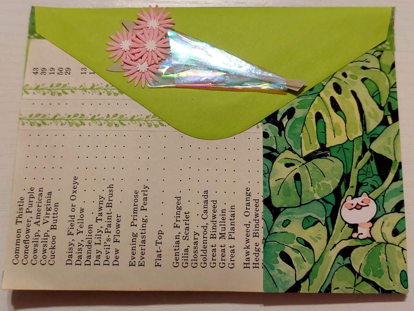 photo of the back of a bright green envelope. the envelope back has a small paper bouquet of flowers pasted onto the envelope flap. there is a page from the directory of a vintage book of flowers turned ninety degrees that is covering half of the bottom of the envelope. the other half has an illustration of monstera leaves and a small white creature sitting on a leaf and smiling at the viewer.