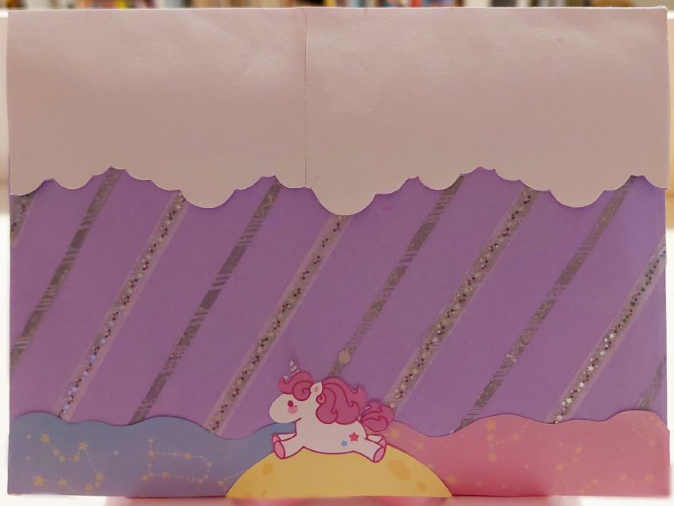 photo of the back of a lavender envelope. there are white clouds on the top flap, diagonal silver stripes on the body of the envelope, and a cartoon unicorn centered at the base of the envelope