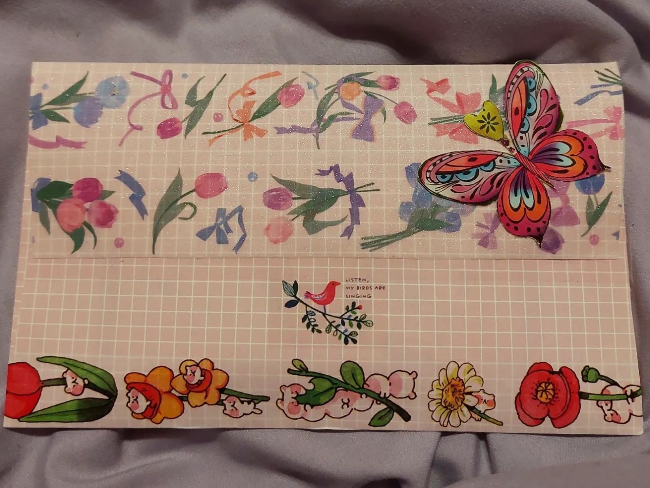 photo of the back of a light pink envelope with a white grid pattern. there are multiple strips of floral washi tape on it and a large, colorful butterfly in the upper right corner.