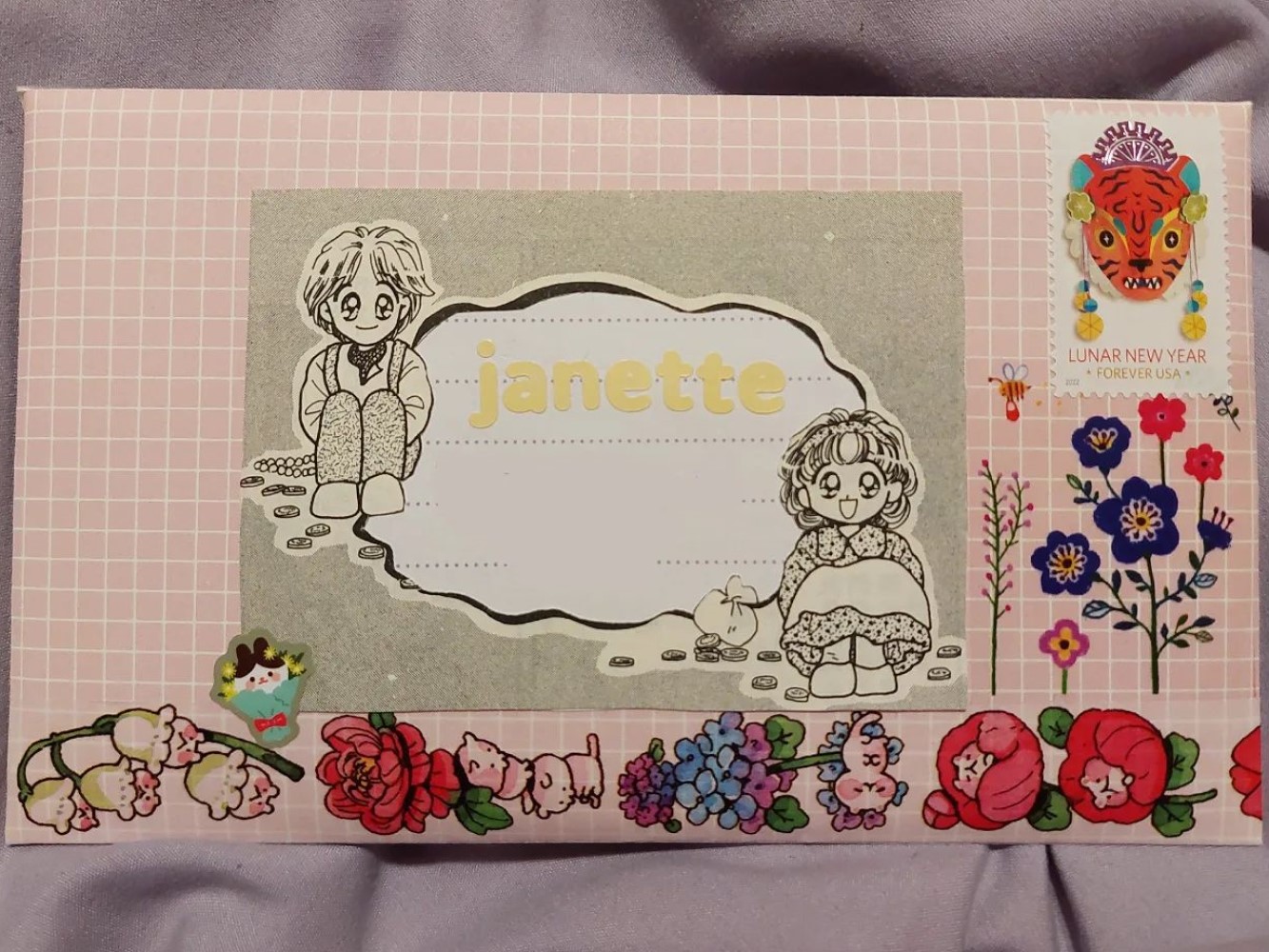 photo of the front of a light pink envelope with a white grid. there is a page cutout from a manga that has two characters sitting opposite each other. between them is a wobbly white shape with the name 'janette' on it in light yellow stickers. colorful flower washi tape decorates the bottom of the envelope