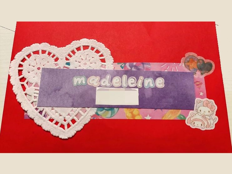 photo of the front of a red envelope. there is a white lace doily in the shape of a heart behind a purple watercolor rectangle with the name 'madeleine' on it. a sticker of my melody and hello kitty in kitty outfits is in the bottom right corner.