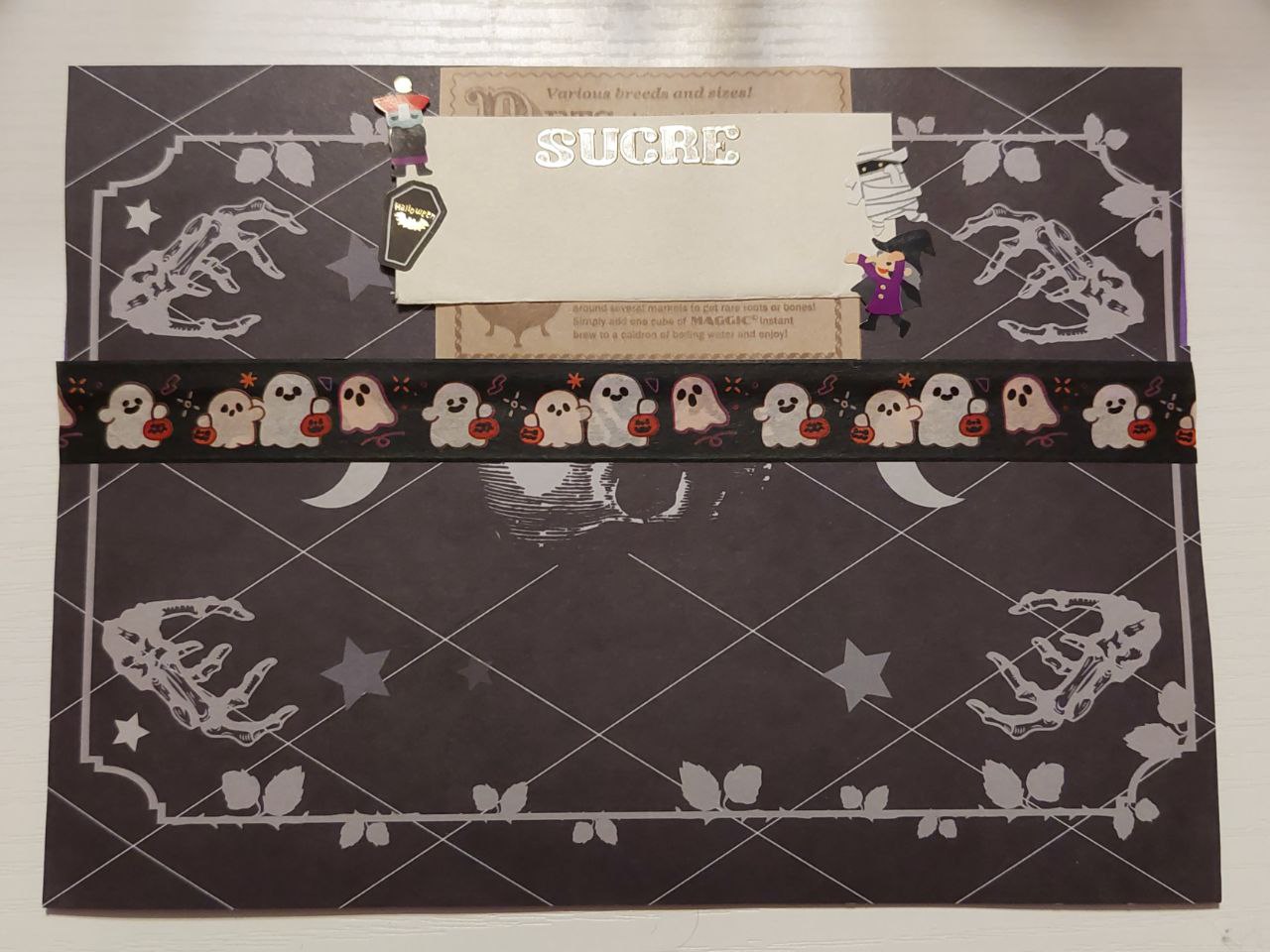 a photo of the back of a envelope. there is black paper with intricate designs pasted onto the envelope. a rectangle at the top has the name SUCRE on it in clear letters with gold edges. there are cute halloween stickers surrounding the rectangle and ghost washi tape sealing the envelope shut.