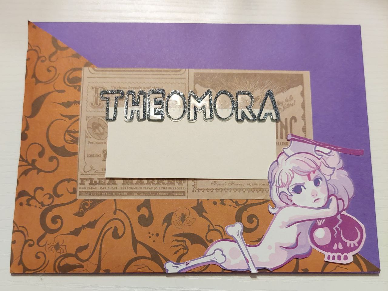 a photo of the front of a purple envelope. there is an orange and black wallpaper talking up half of the letter with an illustration of a woman in the bottom corner. the woman is resting her arms on a skull and has an axe in her head. in the center of the envelope is a rectangle with THEOMORA on it in black glitter letters